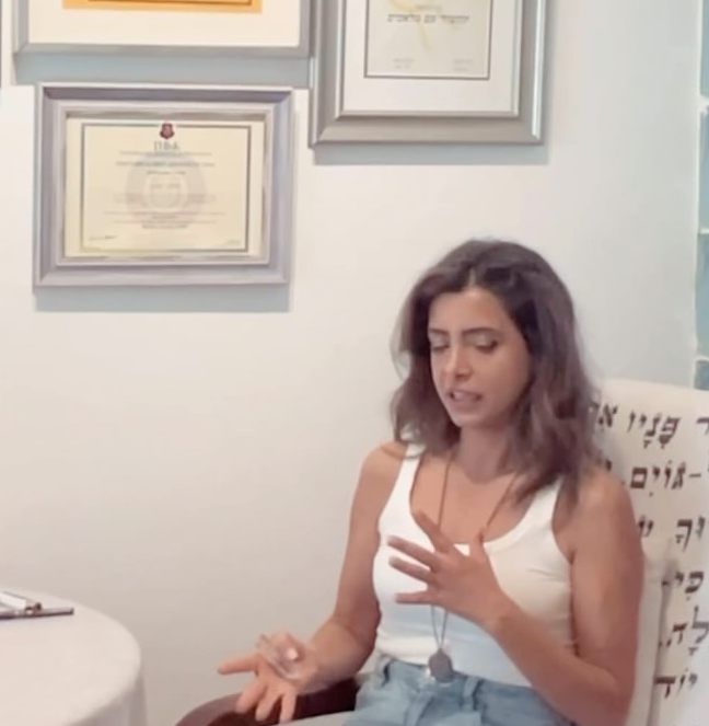 ליאת יושבת בכורסה בקליניקה, ומאחוריה הקיר עם התעודות